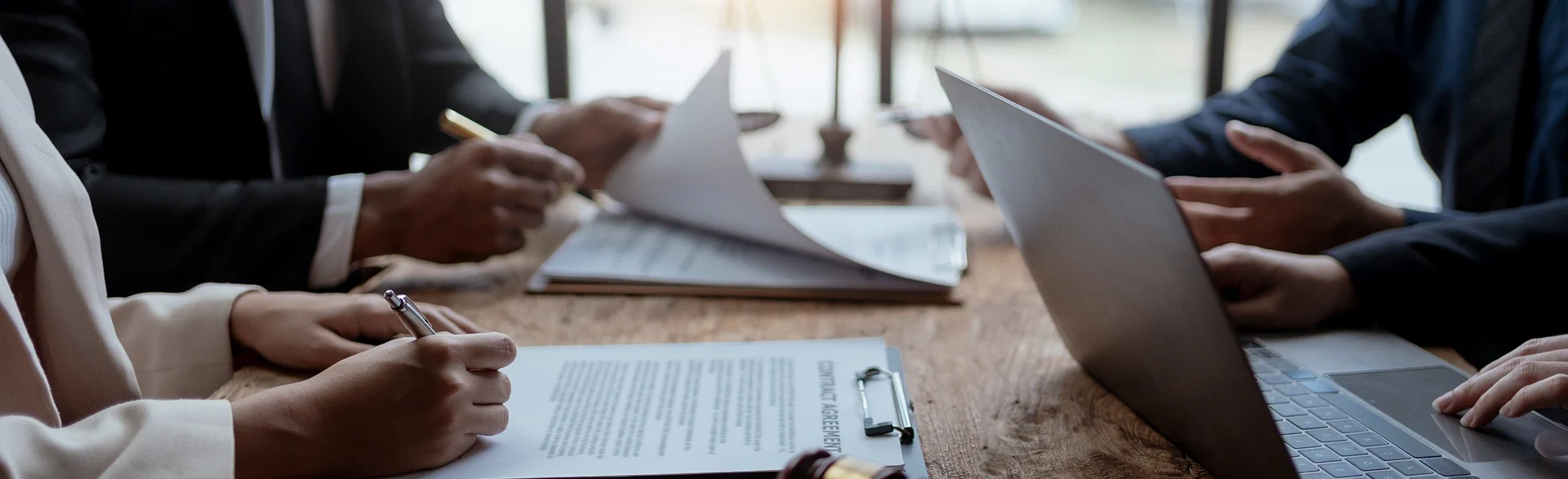 Three people in business attire sit at a table with documents, a laptop, and a gavel, working together in a well-lit office setting