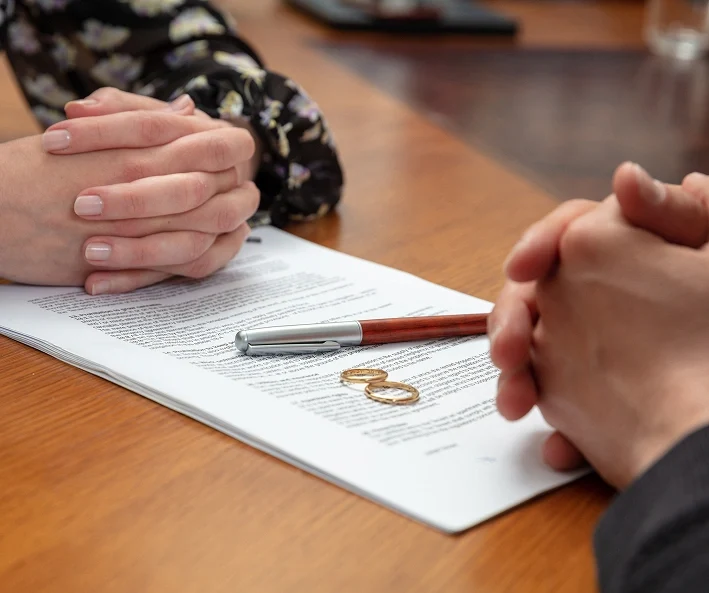 Two people sitting across from each other at a table with a legal document, pen, and two wedding rings placed on top